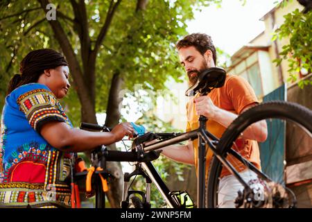 Sana afroamericana signora che assiste il suo ragazzo caucasico riparare la sua bicicletta fuori. Giovane donna nera attiva che fissa la bicicletta al supporto di riparazione per l'atletico da riparare utilizzando attrezzi esperti. Foto Stock