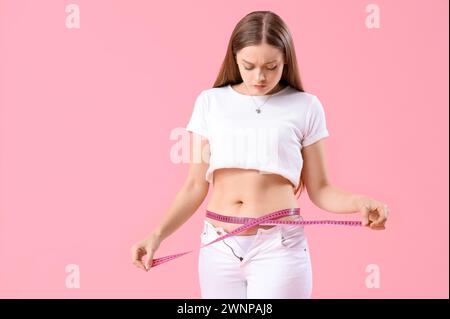 Giovane donna con pantaloni stretti che misura la sua pancia su sfondo rosa. Concetto di aumento di peso Foto Stock
