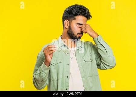 Stanco, stanco, giovane indiano prende gli occhiali, sente dolore agli occhi, essere sovraccarico di lavoro bruciato da lunghe ore di lavoro. Un ragazzo arabo assonnato e stanco che sfrega gli occhi isolato su sfondo giallo Foto Stock