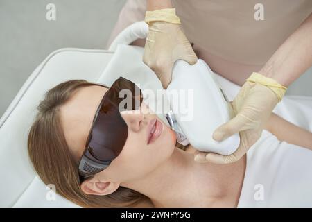 Trattamento viso laser per la depilazione e il ringiovanimento presso la spa. Le attrezzature bianche migliorano la bellezza e la cura della pelle. Il cosmetologo utilizza la tecnologia avanzata per la terapia, promuovendo la salute, la cura cosmetica Foto Stock