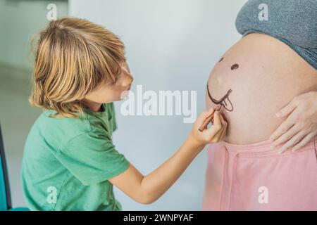 Il momento adorabile di un figlio aggiunge un tocco di gioia alla gravidanza di sua madre, disegnando giocosamente un volto divertente sul batto del suo bambino, creando ricordi preziosi Foto Stock