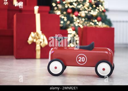 Vista dell'auto retrò rossa per bambini con il numero 20 sullo sfondo dei regali in scatole rosse con archi dorati, ghirlanda decorata con alberi di Natale, luci a corde, palline rosse e blu. Copia spazio, bokeh. Foto Stock