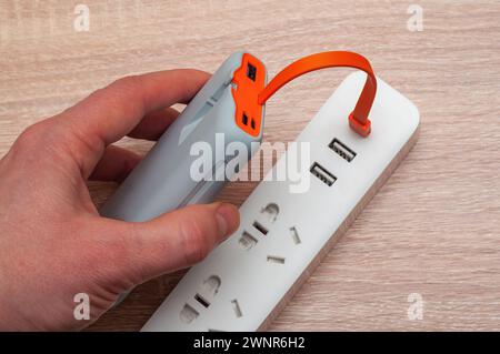 Una mano che regge un power Bank grigio collegato tramite un cavo USB arancione a una prolunga multi-socket bianca con varie porte. Foto Stock