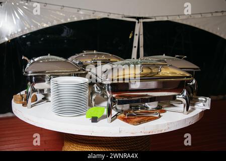 Elegante disposizione a buffet con piatti in acciaio inox e piatti bianchi impilati pronti per un evento con catering. Foto Stock