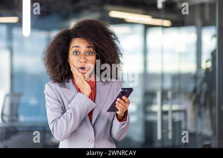 Ritratto di una giovane donna afroamericana scioccata in una tuta da lavoro in piedi in ufficio, tenendo il telefono cellulare, guardando la fotocamera con sorpresa, tenendo mano a faccia. Foto Stock