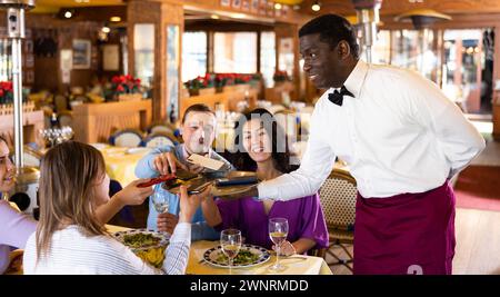 Adulti positivi che danno i loro telefoni al cameriere afroamericano nel ristorante Foto Stock