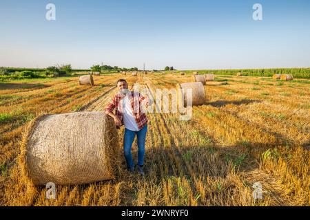 L'agricoltore si trova accanto alle balle di fieno. È soddisfatto dopo aver raccolto con successo. Foto Stock