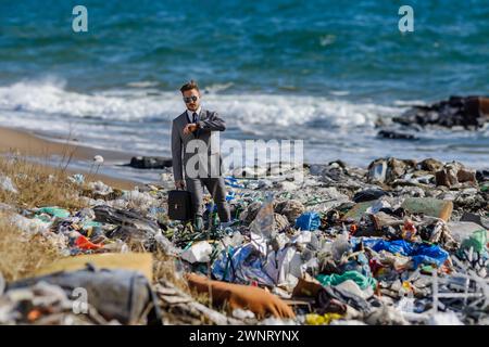 Uomo d'affari che guarda l'orologio, controlla il tempo in piedi su un mucchio di rifiuti sulla spiaggia. Concetto di consumismo contro inquinamento. Responsabilità sociale aziendale Foto Stock