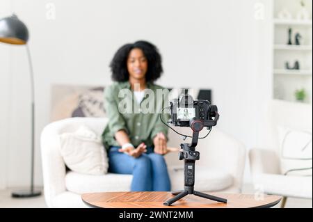 Con un'onda e un sorriso, una donna afroamericana coinvolge il suo pubblico online, creando un'atmosfera invitante nel suo vlog da uno spazio abitativo minimalista Foto Stock