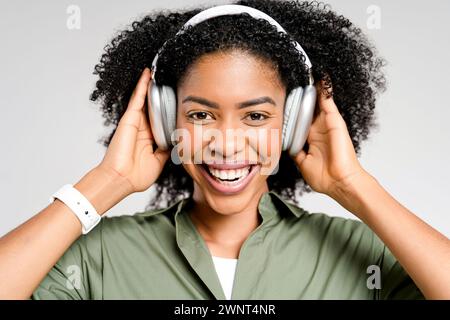 Una allegra donna afro-americana ama la musica preferita con le cuffie color argento, il suo sorriso trasuda la gioia della melodia. Lo studio-shot cattura l'essenza del tempo libero moderno e dell'intrattenimento digitale Foto Stock