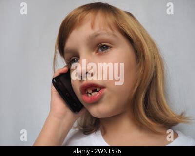 Una bambina di 7 anni senza un dente anteriore. Il bambino sorride, guarda di lato e parla al telefono o allo smartphone. Cambio dei denti del latte Foto Stock