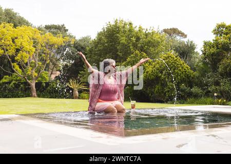 Una donna birazziale senior si tuffa gioiosamente in piscina, beve in mano, in mezzo al verde. Foto Stock