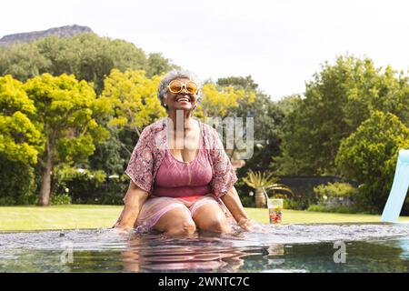 Una donna birazziale senior gode di una giornata di sole in piscina, indossando un top floreale e occhiali da sole Foto Stock