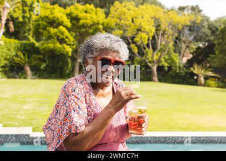 La donna birazziale senior si gode un drink in piscina, mentre i suoi capelli grigi completano il suo abbigliamento vivace Foto Stock