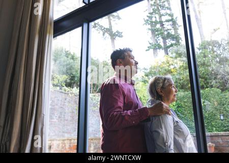 La coppia birazziale senior si trova accanto a una grande finestra, guardando all'esterno con attenzione Foto Stock