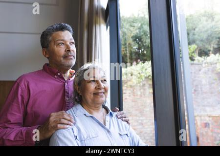 La coppia birazziale condivide un momento tranquillo vicino a una finestra, riflettendo e sorridendo. Foto Stock