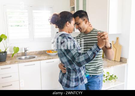 La coppia birazziale gay condivide un momento tenero, ballando in una cucina luminosa Foto Stock