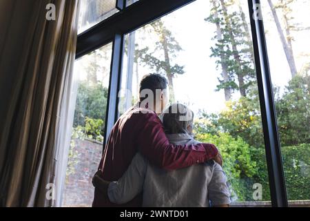 La coppia birazziale senior gode di una vista serena attraverso una grande finestra Foto Stock