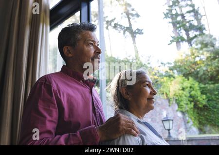 La coppia birazziale senior guarda fuori da una finestra, la donna con striature di grigio nei capelli Foto Stock