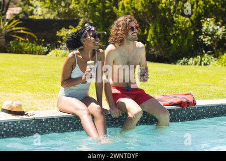 Coppie diverse si godono un drink a bordo piscina, risate evidenti nella loro soleggiata disposizione Foto Stock