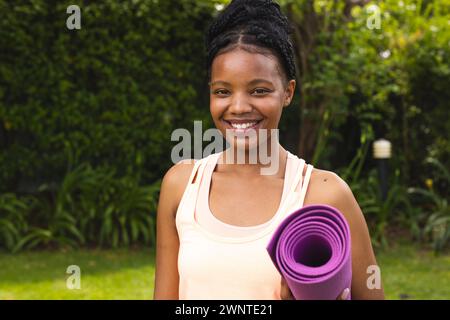 La giovane donna afroamericana con i capelli intrecciati tiene un tappetino yoga viola, sorridendo in un giardino Foto Stock