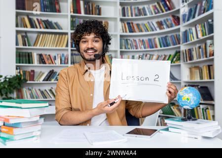 Un giovane e coinvolgente insegnante di sesso maschile sorride mentre presenta "lezione 2" su una lavagna, circondato da pile di libri in un luminoso ambiente bibliotecario che simboleggia l'educazione Foto Stock