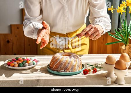 Le mani femminili cospargono la torta di Pasqua con zucchero a velo attraverso il setaccio. Donna anonima che prepara la tradizionale torta di pasqua o il pane dolce con condimento. Pasqua Foto Stock