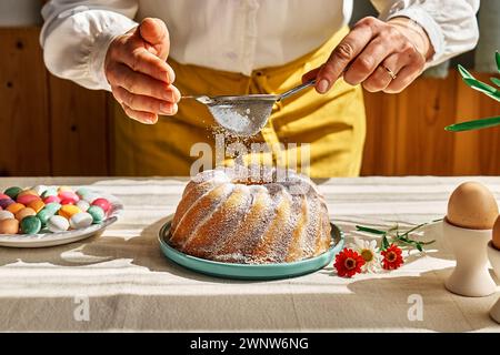 Le mani femminili cospargono la torta di Pasqua con zucchero a velo attraverso il setaccio. Donna anonima che prepara la tradizionale torta di pasqua o il pane dolce con condimento. Pasqua Foto Stock