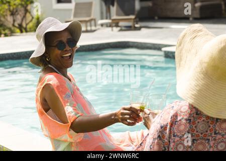 Una donna afroamericana anziana in un cappello da sole ride con una donna birazziale in piscina, con spazio per le copie Foto Stock