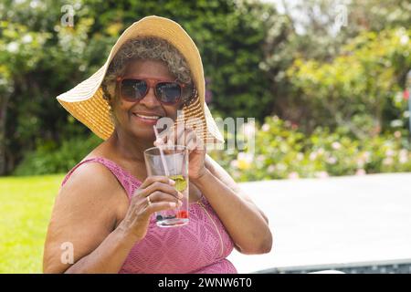 Una donna birazziale senior gode di un drink all'aperto con spazio per le copie, indossando un cappello da sole e occhiali da sole Foto Stock