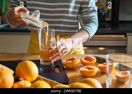 Mani femminili che preparano il succo di pompelmo fatto in casa con la centrifuga di agrumi a mano. Donna anonima che versa succo di pompelmo fresco in un bicchiere in cucina Foto Stock