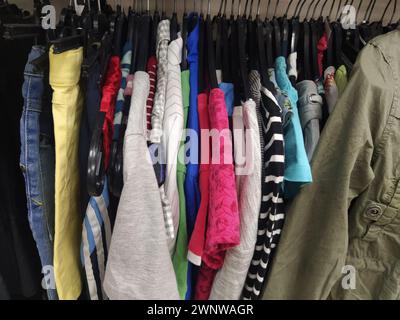 Gli abiti sono appesi alle grucce. Assortimento di articoli usati. Camicie, camicette, maglioni da donna e bambini appesi a un rack, pronti per la vendita a. Foto Stock