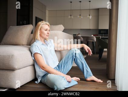 Ritratto di donna seduta accanto al divano nel suo appartamento. Bella donna in abiti casual che guarda lontano. Foto Stock