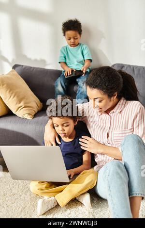 gioiosa donna afro-americana che passa del tempo con i suoi due figli simpatici che reggono laptop e telefono Foto Stock