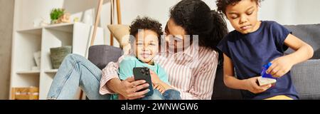 allegra madre afroamericana con il telefono in mano, trascorrendo del tempo con i suoi due adorabili figli, striscione Foto Stock