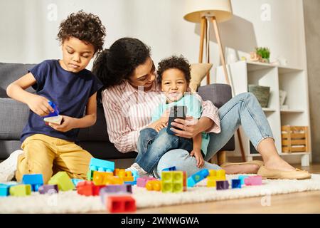 gioiosa madre afroamericana con il telefono in mano, trascorrendo del tempo con i suoi due adorabili figli Foto Stock