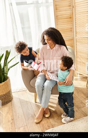 Felice attraente donna afroamericana che riceve cartolina e regalo dai suoi figli, festa della mamma Foto Stock