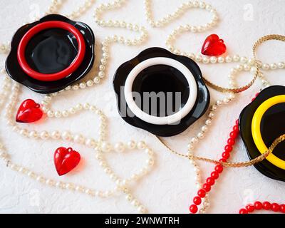 Posizione piatta. Una splendida composizione di piatti neri in vetro, gioielli in plastica, bracciali, perline, cuori rossi su sfondo bianco. Baubles femminili per Foto Stock