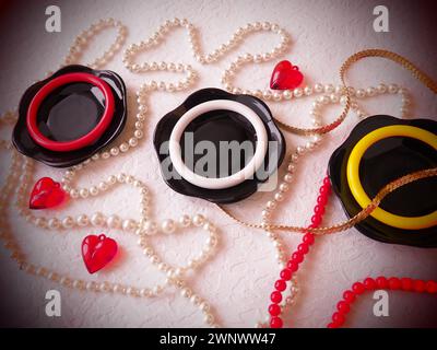Posizione piatta. Una splendida composizione di piatti neri in vetro, gioielli in plastica, bracciali, perline, cuori rossi su sfondo bianco. Baubles femminili per Foto Stock
