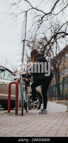 Una donna con abbigliamento attivo mette in sicurezza la sua moto su una strada cittadina, mostrando uno stile di vita urbano e la routine quotidiana dei ciclisti. Foto Stock