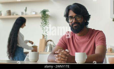 Felice barbuto uomo proprietario di casa marito sedersi in cucina musulmano indiano ragazzo sorridente allegro sorriso mentre donna moglie cucinare colazione lavoro domestico cucina Foto Stock