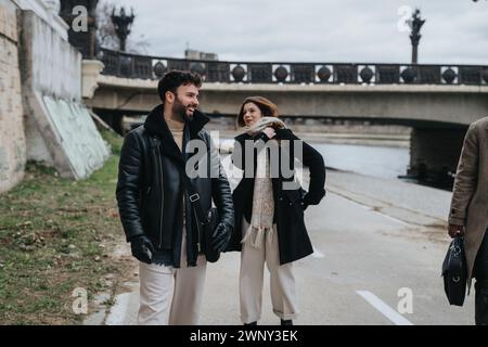 Giovane coppia gioiosa che si gode una passeggiata lungo il fiume in città, immergendosi in una vivace conversazione con cappotti invernali. Foto Stock