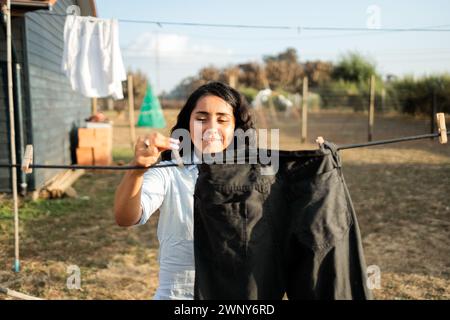 Modest Domestic Scene: La giovane donna latina si occupa di lavanderia asciugando i vestiti nel cortile di campagna. Concetto di lavoro domestico Foto Stock