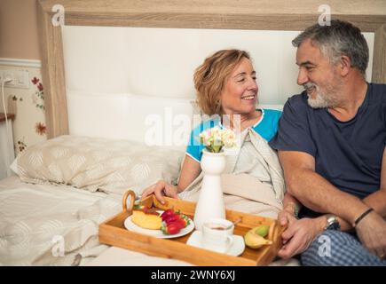 Questa splendida immagine cattura un momento intimo tra una coppia di anziani che condivide la colazione a letto. Con uno sguardo pieno di amore e intimità, loro d Foto Stock