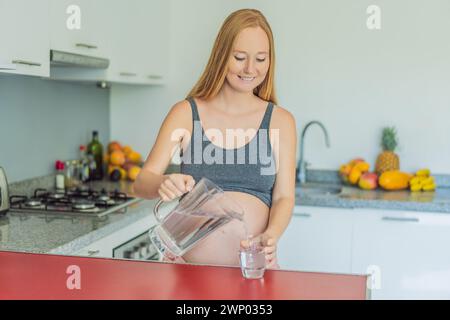 Abbracciando i benefici vitali dell'acqua durante la gravidanza, una donna incinta sta in cucina con un bicchiere, evidenziando il ruolo cruciale dell'idratazione in Foto Stock