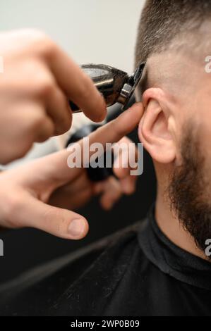 Il barbiere rasa il contorno della linea ovale con un regolacapelli sulla testa del cliente. Un uomo con la barba si fa tagliare i capelli su una sedia da barbiere. Foto Stock