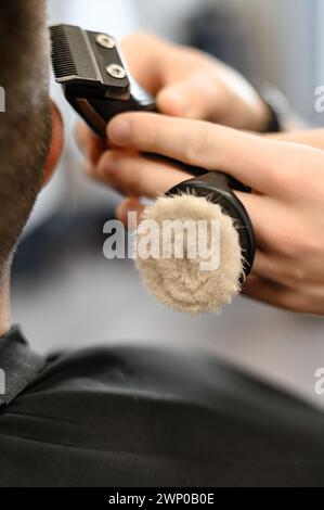 Il barbiere rasa il contorno della linea ovale con un regolacapelli sulla testa del cliente. Un uomo con la barba si fa tagliare i capelli su una sedia da barbiere. Foto Stock