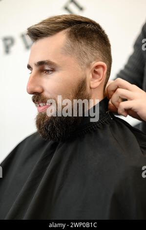 Un giovane con la barba in un mantello nero siede su una sedia in un barbiere. Il cliente sta aspettando un taglio di capelli e la rasatura della barba. Foto Stock