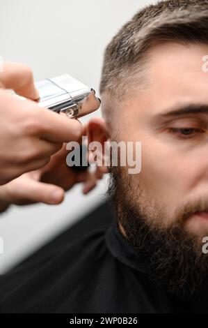 Il barbiere rade la stanghetta con un rifinitore cordless durante un breve taglio di capelli ai lati della testina. Foto Stock