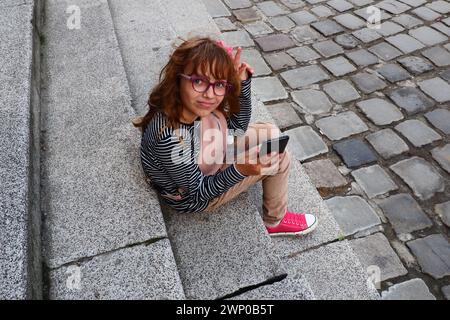 Una bambina di 8 anni che indossa gli occhiali si siede sulle scale di una chiesa a Petrovaradin, guarda e usa un telefono o uno smartphone. Vita cittadina e.. Foto Stock